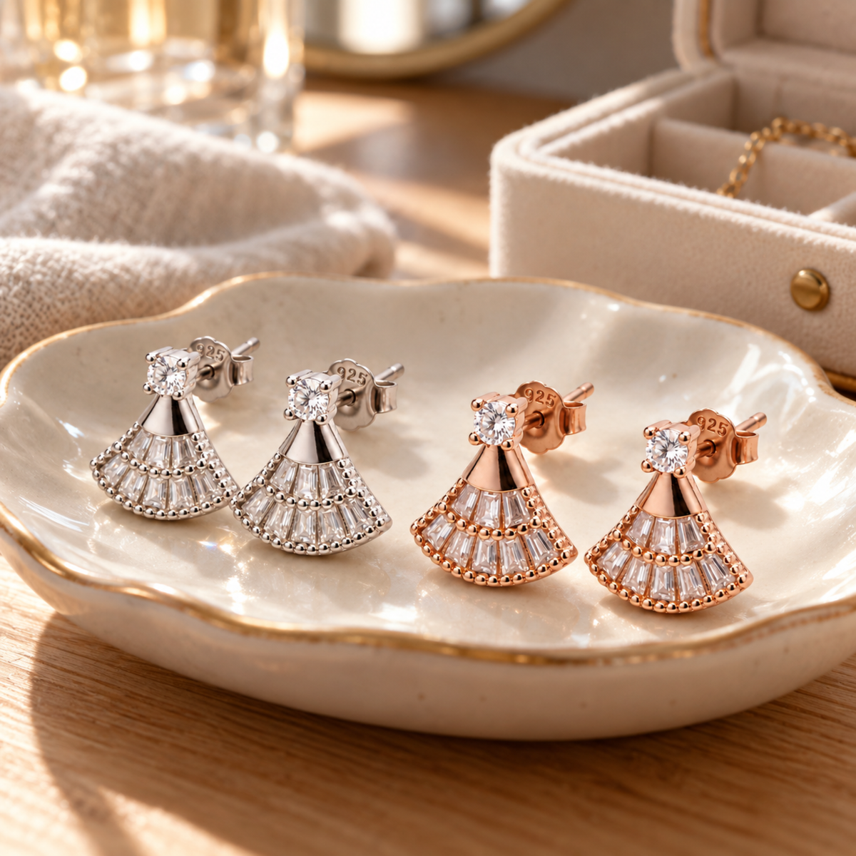 Four drop earrings displayed on a scalloped cream dish, featuring silver and rose gold metal settings with clear baguette and round-cut stones. The dish sits on a wooden table with a soft beige towel and a cream jewelry box in the background.