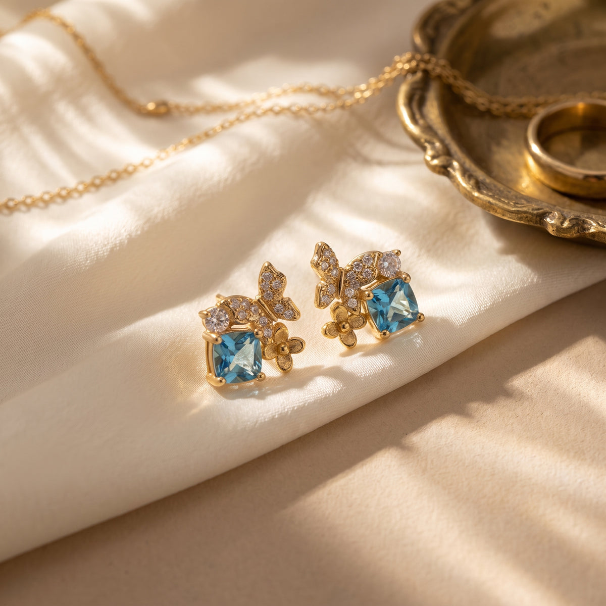 Blue Gold Butterfly Garden stud earrings displayed on soft champagne satin