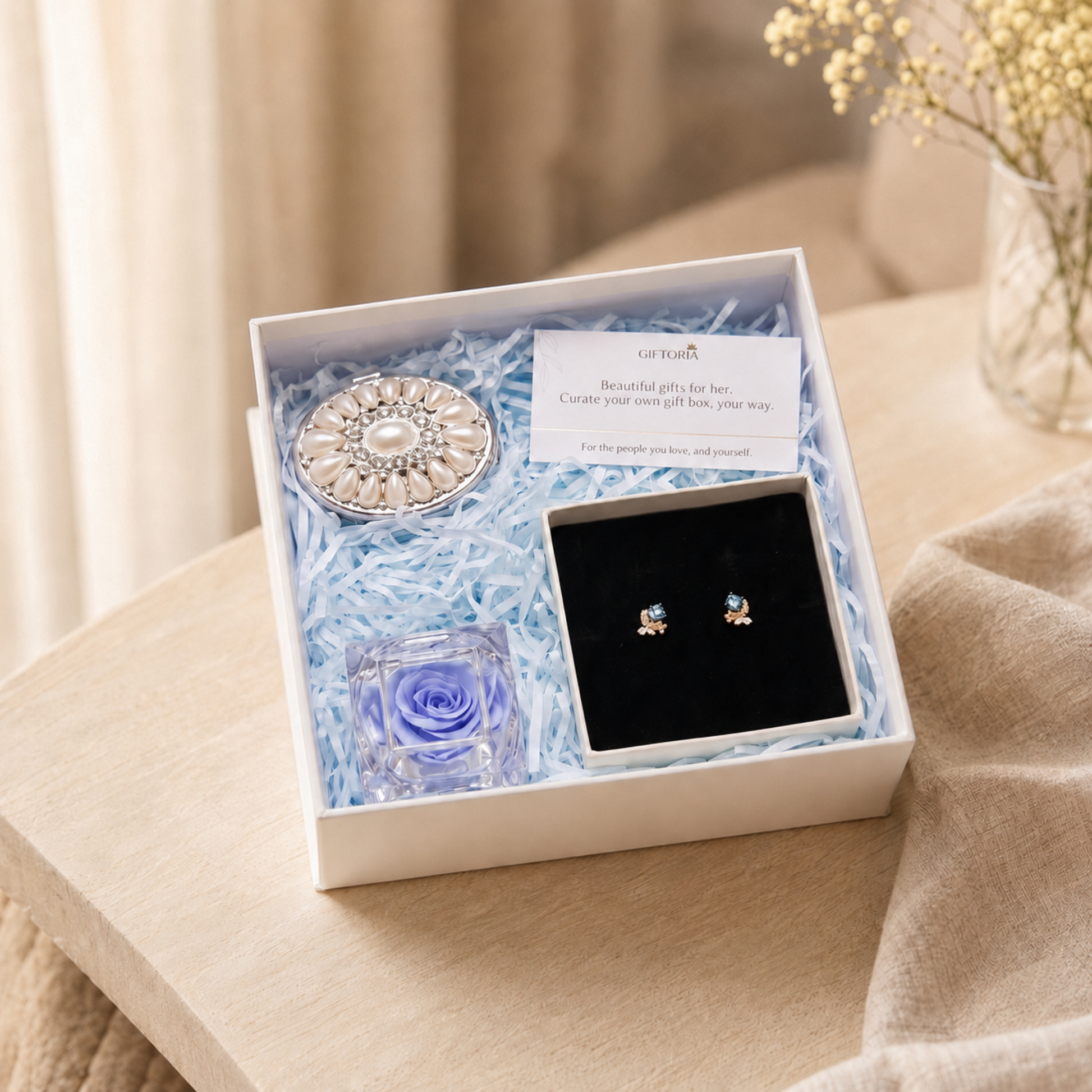 Gold butterfly earrings, blue preserved rose and pearl jewellery box gift set on a beige fabric surface, featuring a round silver-toned brooch with a central pearl, a small square glass vial with a blue rose, a black jewelry tray holding a pair of small gold and blue stud earrings, and a white card. The box is filled with light blue shredded paper.