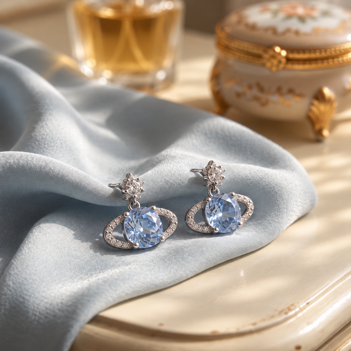 Blue Rose Saturn silver stud earrings styled on blue satin with gold-toned accessories in the background.