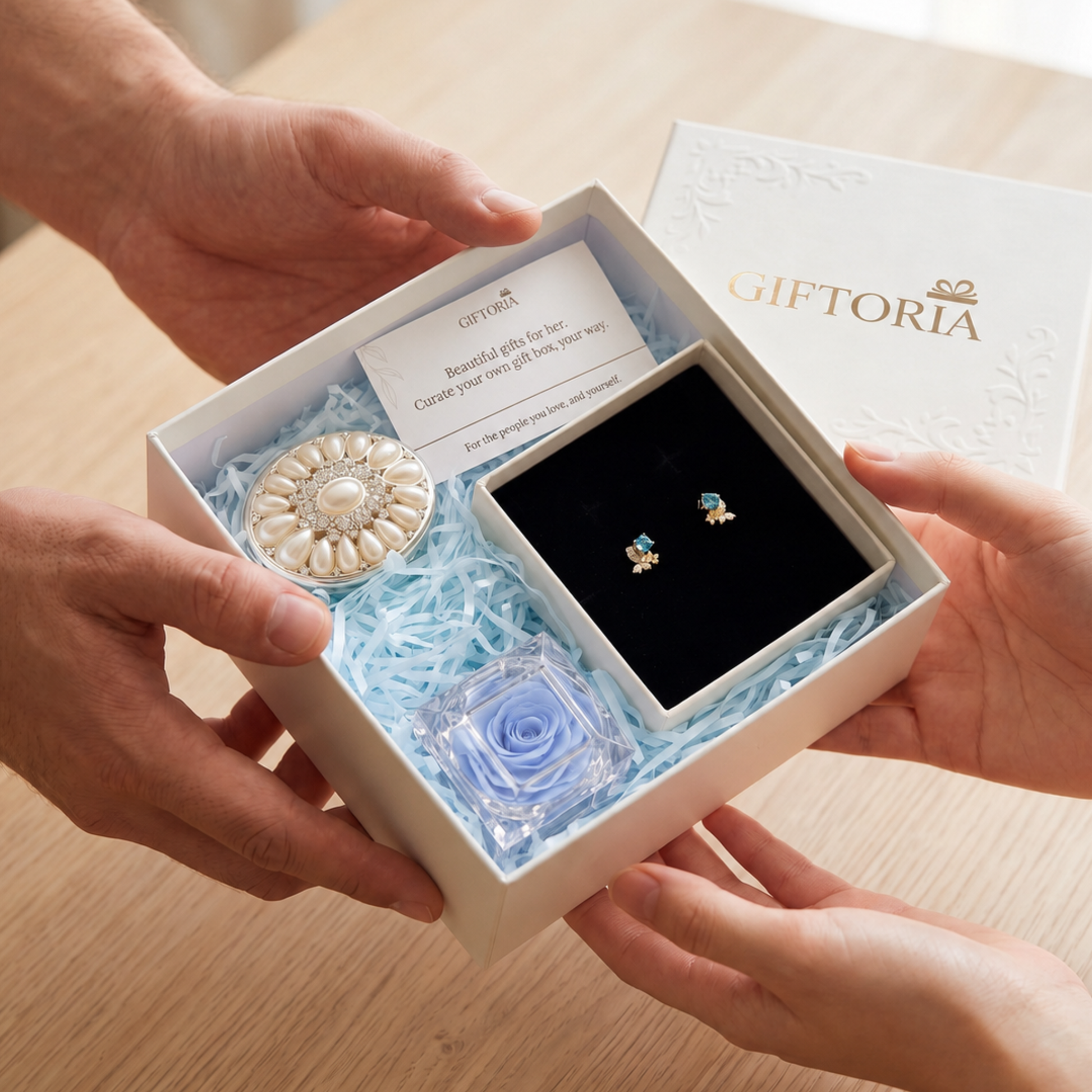 Hands presenting a Giftoria gift box with gold butterfly earrings, blue preserved rose and pearl jewellery box