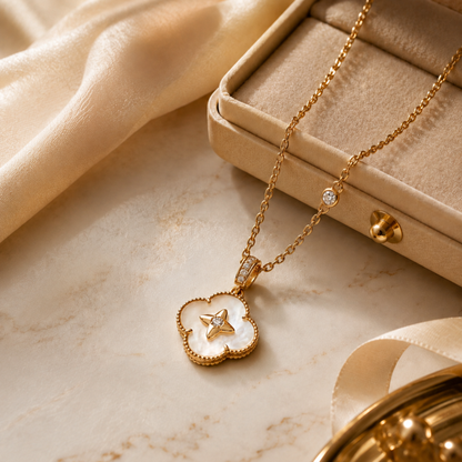 A gold-tone necklace with a clover-shaped pendant featuring a white inlay and a small star motif, styled on a beige marble surface next to a beige jewelry box and draped satin fabric.