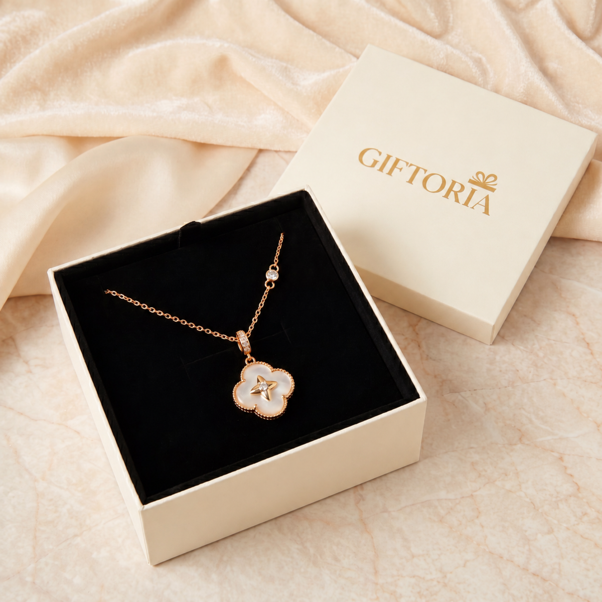 A gold-tone necklace with a white enamel clover pendant is displayed in an open cream-colored gift box with a black velvet interior. The cream-colored brand box lid, printed with the name 'GIFTORIA', rests beside it on a beige marble surface with draped cream satin fabric in the background.