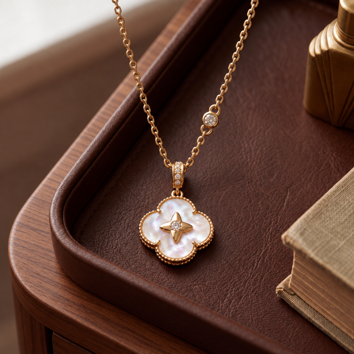 A gold-tone necklace with a mother-of-pearl four-petal pendant and a small round clear stone on the chain, displayed on a brown leather tray with a wooden table and books in the background.