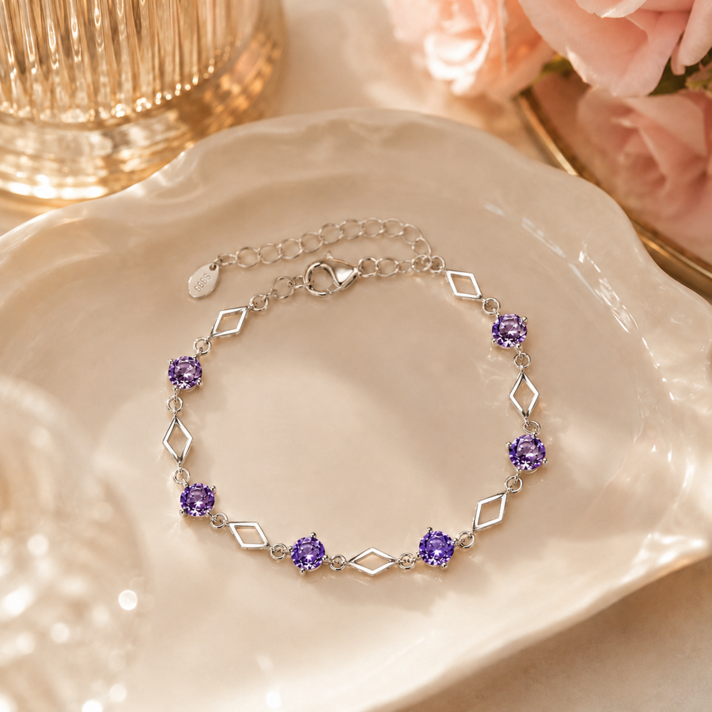 Luminous Love purple cubic zirconia 999 fine silver bracelet displayed on a glossy cream dish with soft pink flowers and a gold-toned glass in the background.