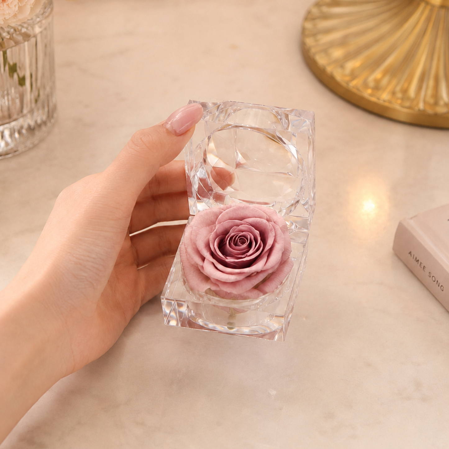 A hand holds a rose gold Preserved Rose Crystal Gift Box with a real preserved rose inside a clear crystal box set on a light marble surface with soft decorative background elements.