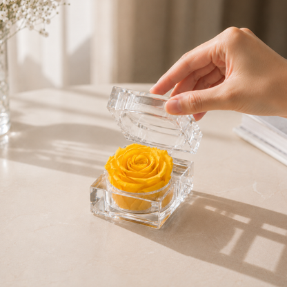 A hand is lifting the yellow Preserved Rose Crystal Gift Box with a real preserved rose inside a clear crystal box, set on a light beige tabletop in warm natural light.