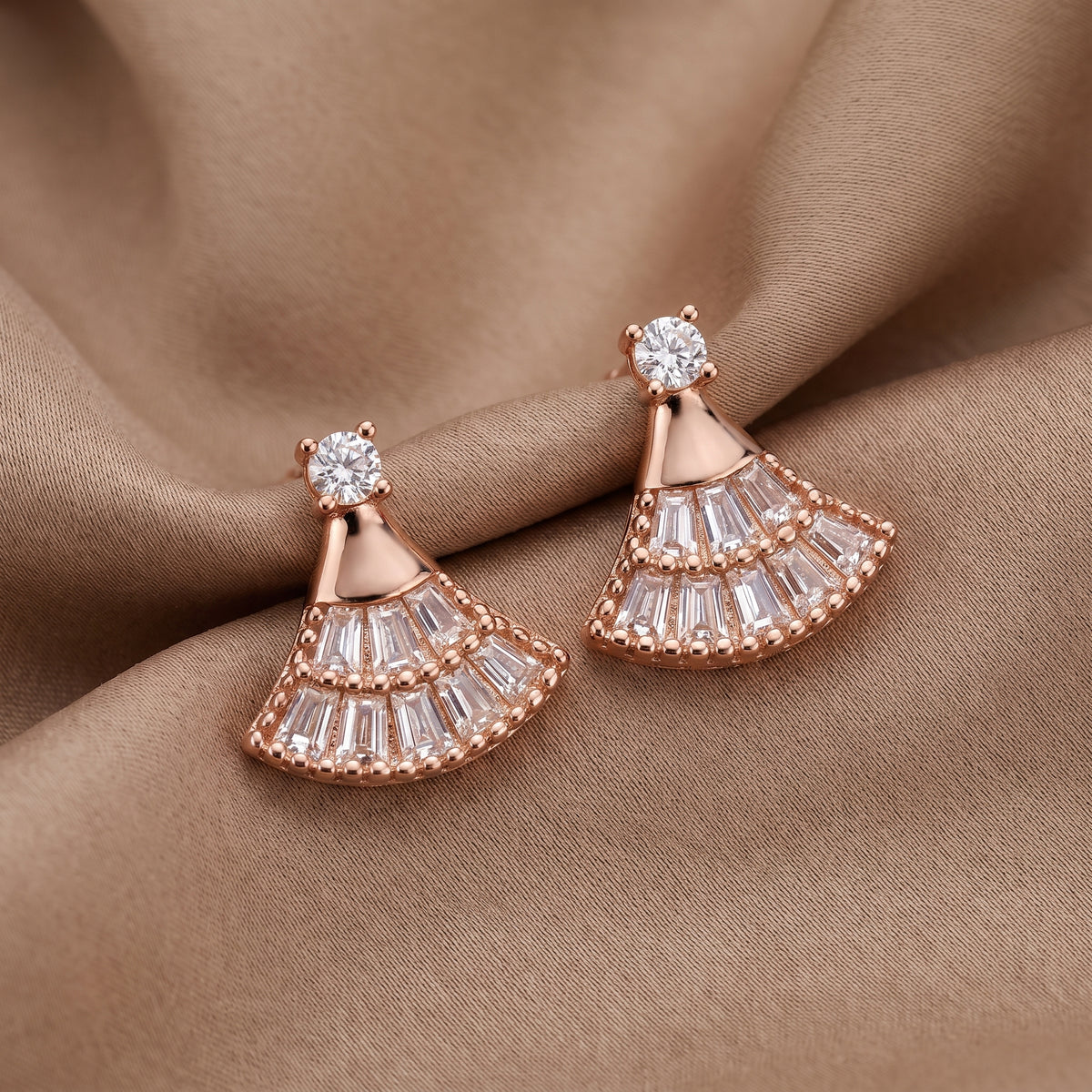 Rose gold fan-shaped drop earrings with clear baguette and round stones, displayed on draped brown fabric.