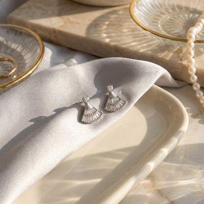 Silver fan-shaped earrings on soft fabric and a cream tray in warm light.